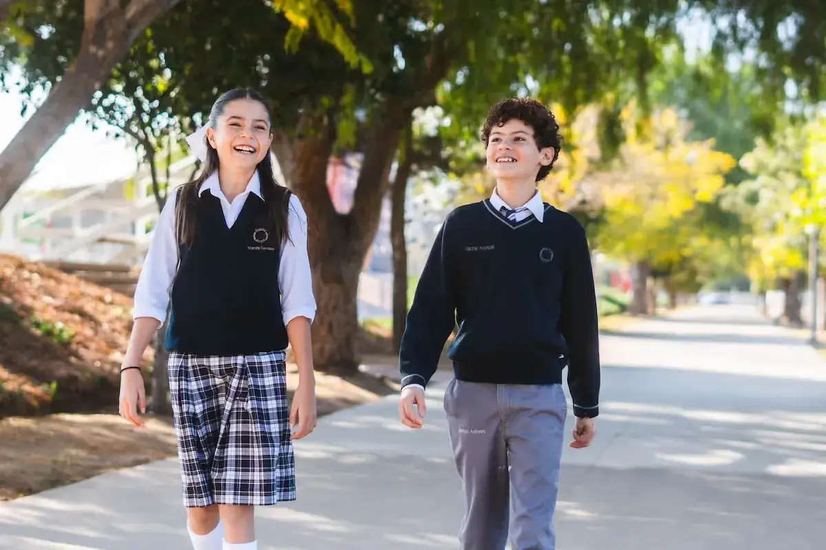 Dos estudiantes con uniforme escolar caminan juntos bajo árboles en el campus durante el día.